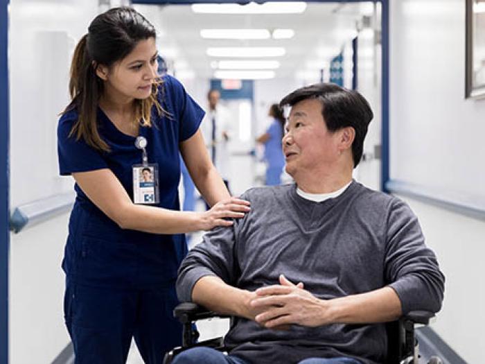 Nurse with dark hair and wearing dark blue scrubswith hand on shoulder of Asian main in wheelchair