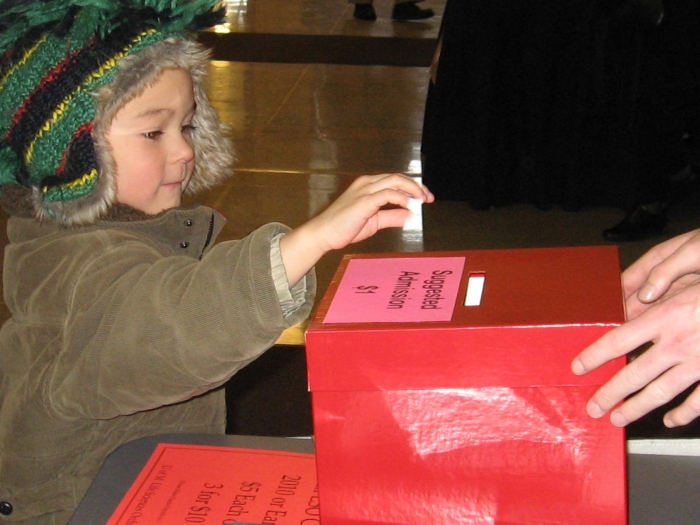 Little boy with striped green knit hat with earflaps putting something in a red donation box