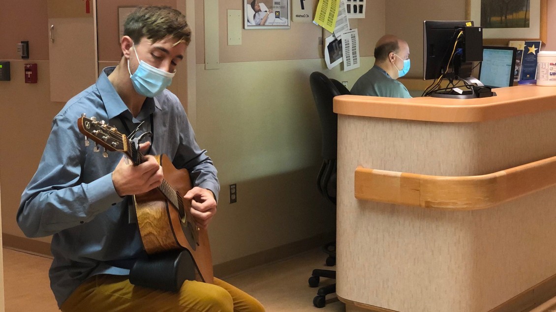 Man wearing mask playing guitar in hospital with receptionist seated in background