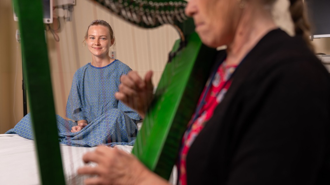 Woman playing green harp in foreground with patient seated on hospital bed in background listening