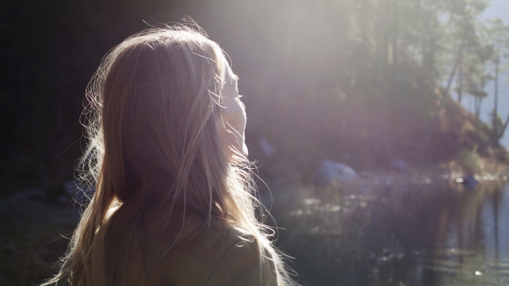 Sunlit woman with long blonde hair gazing into distance at water and trees