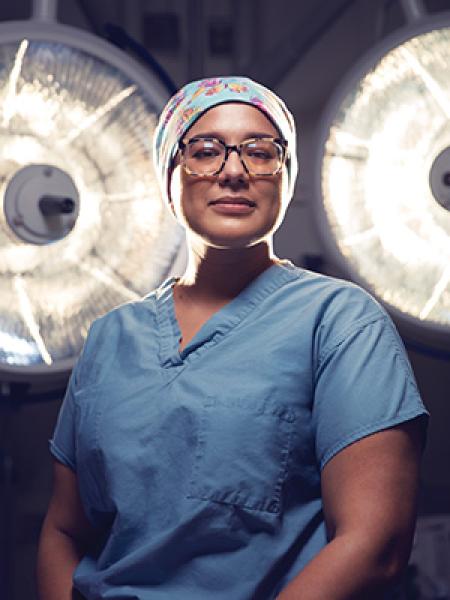Female doctor wearing scrubs and glasses with large surgical lights behind her