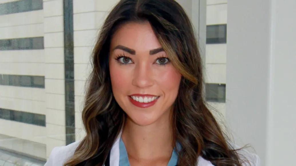 A young Asian American woman with long curled brown hair, wearing blue scrubs, a white doctor's coat, and a stethoscope