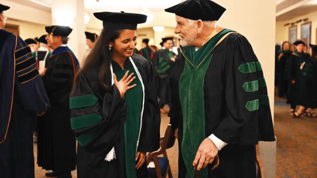 Two people in black and green graduation robes share a happy moment of celebration in a crowded lobby. The woman on the left is younger and smiling or laughing. The older man on the right is carrying a cane and smiling at the woman.