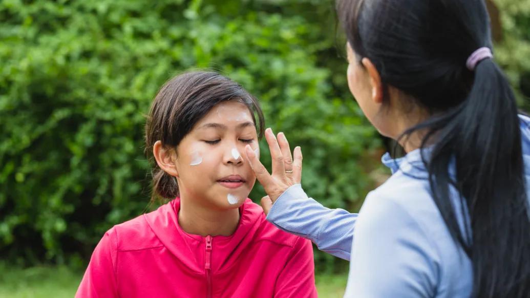 A mom applies sunscreen to her daughter's face. They are outdoors in a green area. Both are wearing long-sleeve jackets.