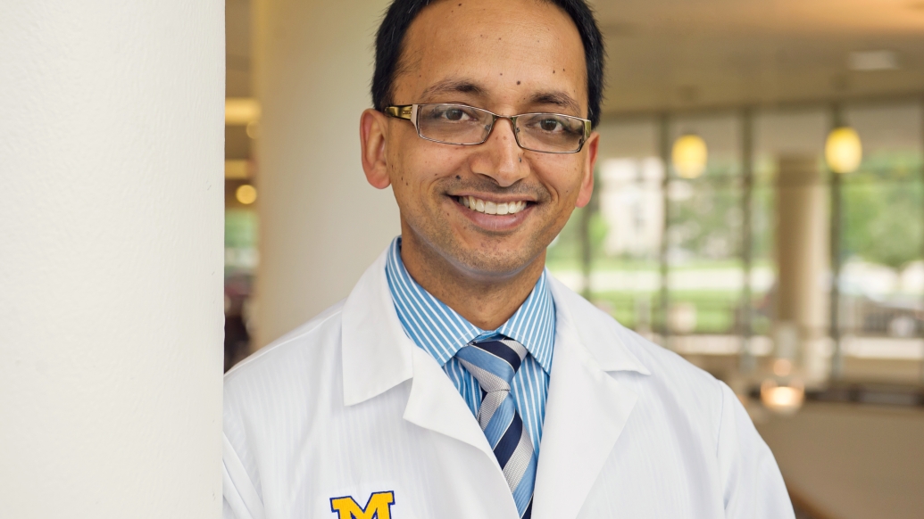 Hitinder Gurm, M.D. wearing a blue shirt, striped tie, and a Michigan Medicine branded white lab coat, standing next to a pillar
