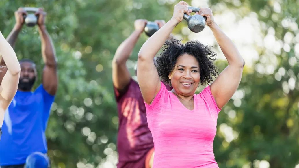 Woman in pink shirt lifts kettleball in an outdoor exercise class