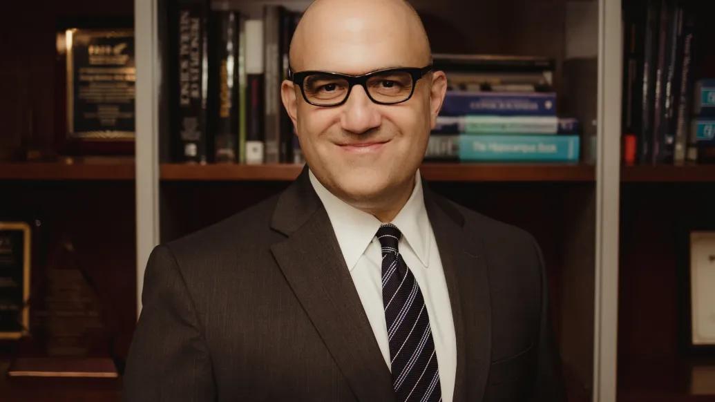 George Mashour, M.D., Ph.D. standing in front of a bookcase, smiling. He is wearing a dark suit, white shirt, dark striped tie and dark framed glasses.