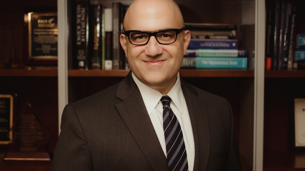 George Mashour, M.D., Ph.D. standing in front of a bookcase, smiling. He is wearing a dark suit, white shirt, dark striped tie and dark framed glasses.