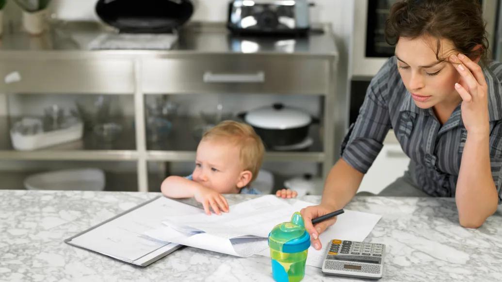 Mom at table with baby, working on calculator with stressed out first expression