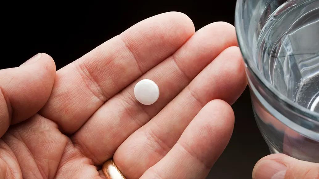 Person's hand holding an aspirin tablet with a glass of water nearby