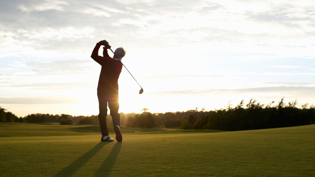 Older man swings a golf club with sunset in the background