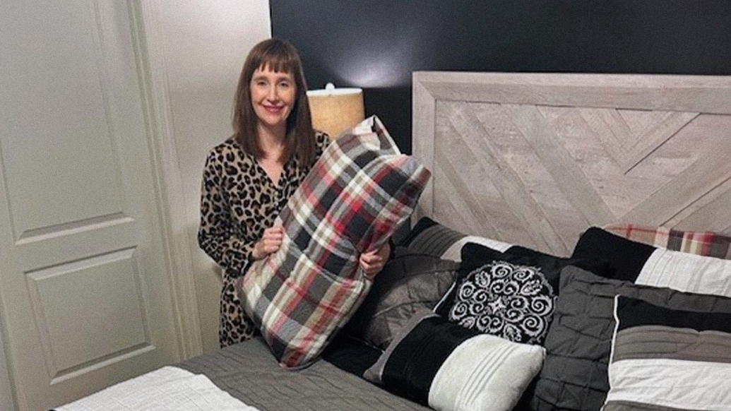 smiling woman holds pillow with plaid cover next to bed