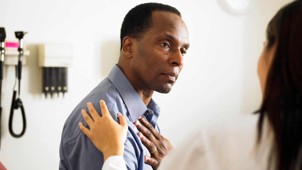 black patient in a doctor's office, with caregiver who has hand placed on his shoulder