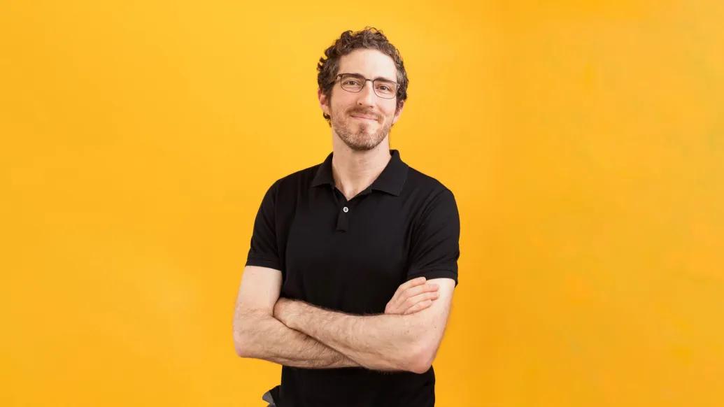 man in black polo folded arms smiling no teeth bright orange background