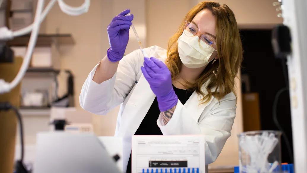 woman putting something in vial in lab coat and purple gloves, glasses and mask in clinical looking area