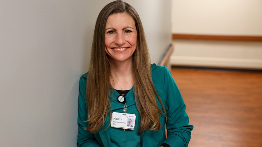 woman with brown long hair and green top and ID badge on smiling at camera against gray wall with brown floor behind and brown thin banisters on wall 