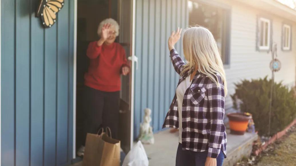 older woman in red long sleeve shirt waving from open door of blue house with other woman leaving with long silver hair and checkered shirt with white undershirt turned towards the door