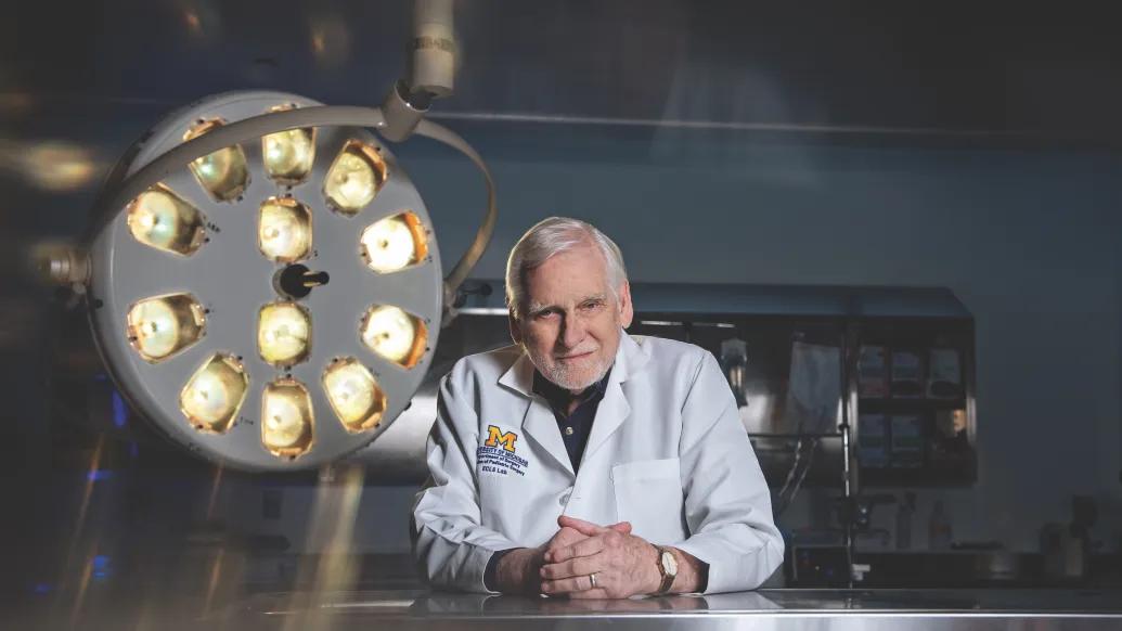 Dr. Robert Bartlett is wearing a doctor's white coat and leaning on a steel table in a lab with his hands clasped. He's standing next to a large circular surgical light. 