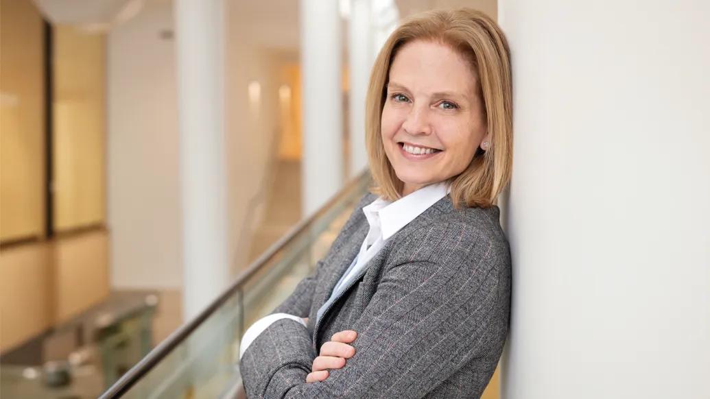 Headshot of Carrie Cunningham. She's wearing a gray suit jacket with a white shirt. She has shoulder-length blond hair and is leaning against a wall with her arms crossed. 