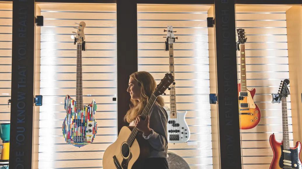 Erica Ginepro stands in front of a wall display of different types of guitars. She's holding an acoustic guitar, is facing sideways, and is looking to the side.