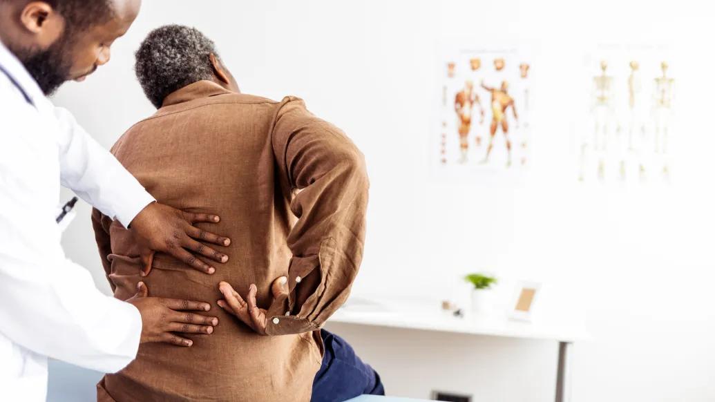 A doctor examines a patient's back. His hand's are on the patient's lower back and the patient is reaching around pointing to a spot on his back. 