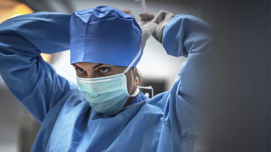 female surgeon in blue cap scrubs and mask tying mask and looking up 