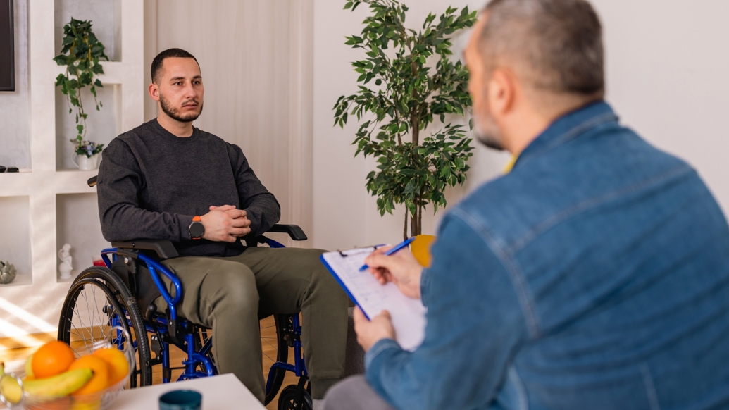 man in wheelchair talking to other man with back to camera in jean shirt in living room looking setting with tree in corner