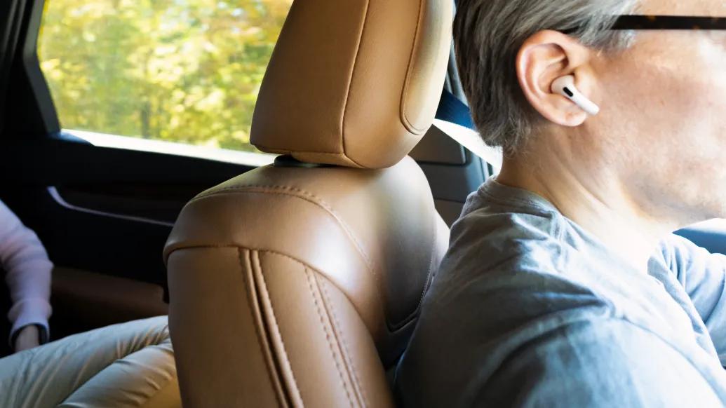 close up of airpod in ear while old man with glasses drives and light brown leather seats in car