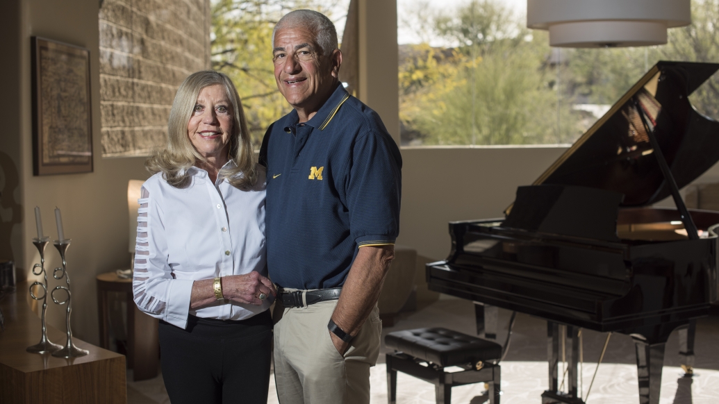 Susan and Richard Rogel pose next to a grand piano. Rogel is wearing a polo shirt with a block M.