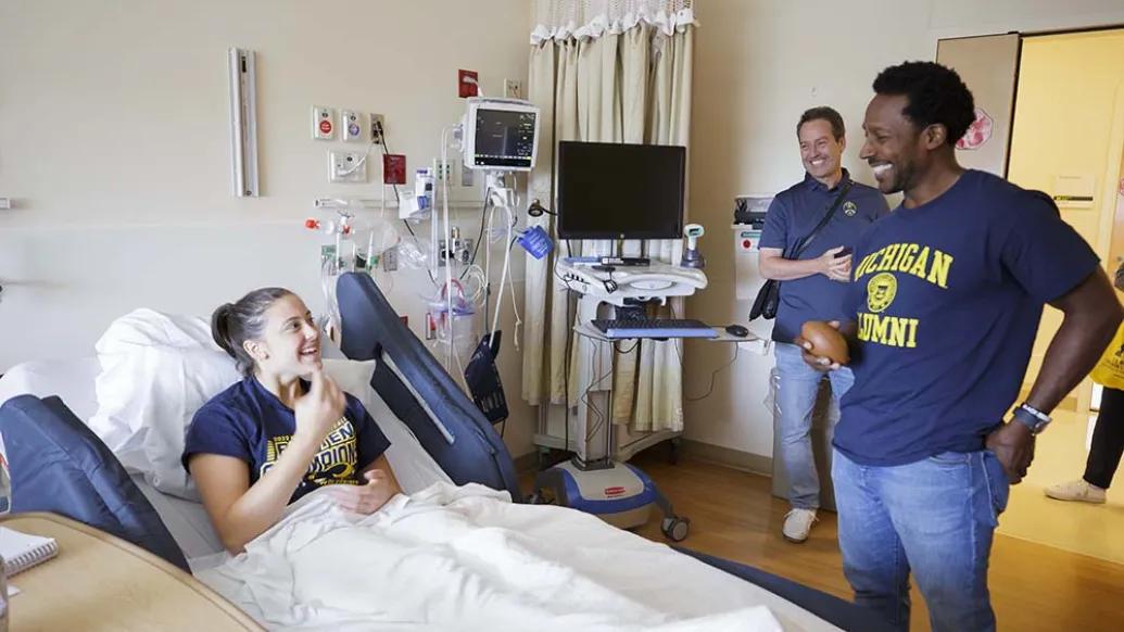 Desmond Howard visits a patient at Mott. He is smiling and wearing a U-M alumni t-shirt and jeans. The patient is lying on a hospital bed and is covered with a white blanket. 