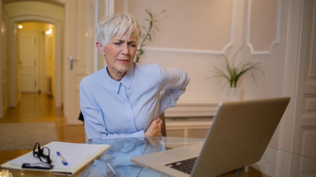 woman touching back leaning forward in pain at desk light powder blue button down