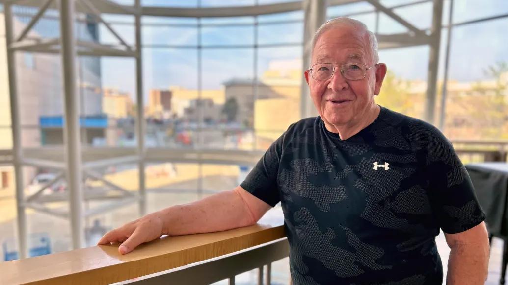 older man with glasses standing at balcony with back to glass windows 