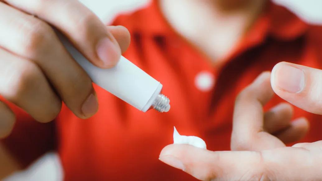 white cream being put on finger close up with blurred red shirt in background of person using cream
