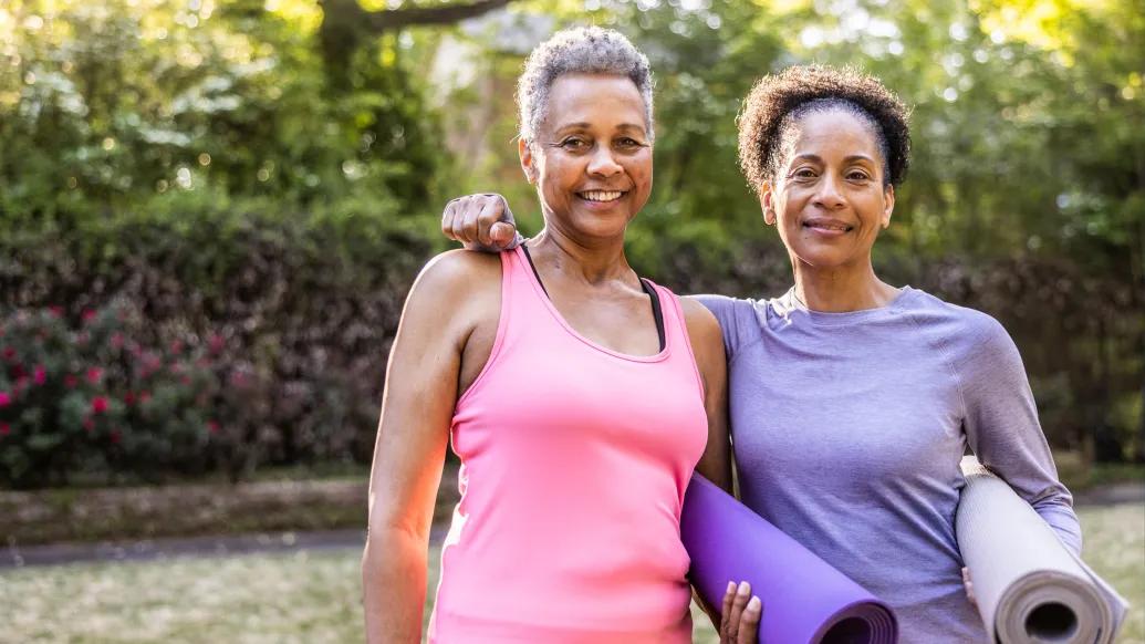 two Black women standing next to eachother, one in a pink tank top, one in long sleeve purple shirt, with yoga purple matts