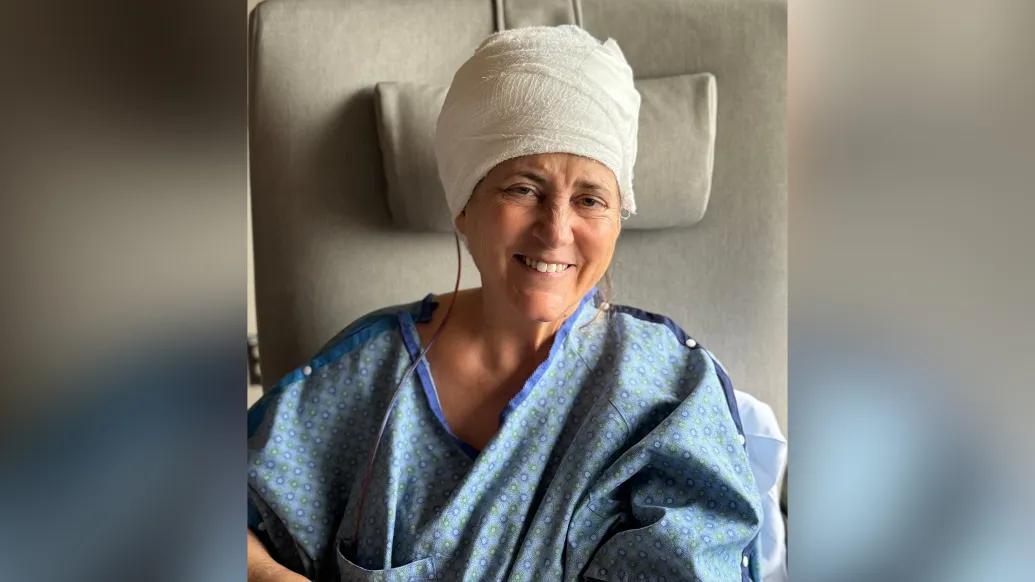 woman smiling after brain surgery with brain wrapped in white cloth and in blue hospital gown on grey background