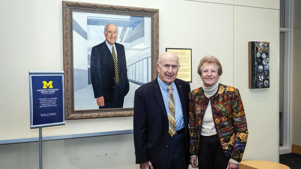 Gilbert Omenn and Martha Darling pose in front of a portrait of Omenn at the University of Michigan Medical School. Omenn is wearing a suit and tie and Darling is wearing a colorful print blazer.