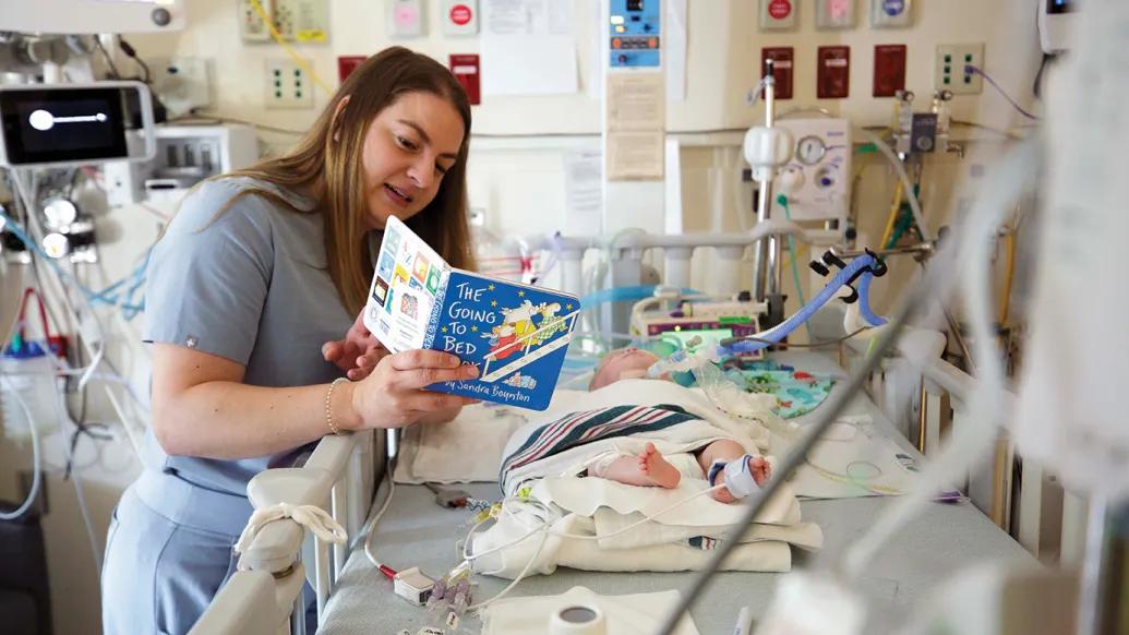 A nurse reads The Going to Bed Book by Sandra Boynton to a baby who is in the Neonatal Intensive Care Unit. The baby is in a hospital crib and hooked up to several machines. The nurse is wearing blue-gray scrubs.