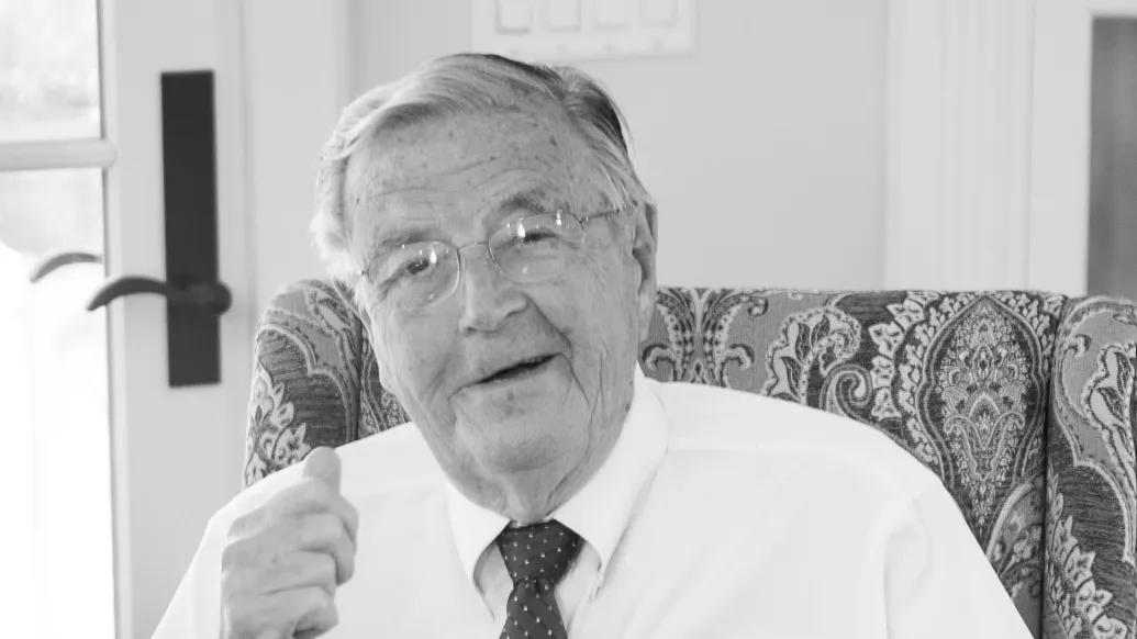Smiling black and white photo of Phil Jenkins seated at his desk. He is dressed up in a button-up shirt and tie.