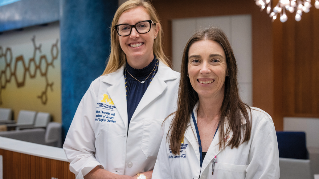 Melissa Pilewskie, M.D., associate professor of surgery, and Monika Burness, M.D., assistant professor of internal medicine, pose together on the Medical Campus. They are both wearing white coats, smiling, and looking at the camera.
