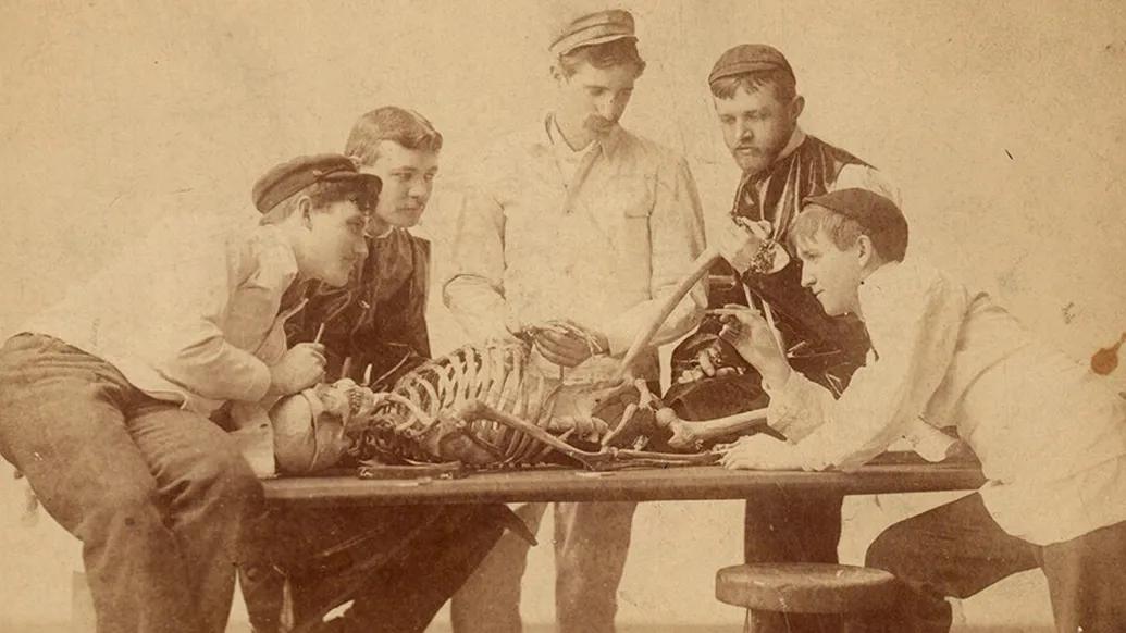 old photo of men sitting around skeleton on table talking