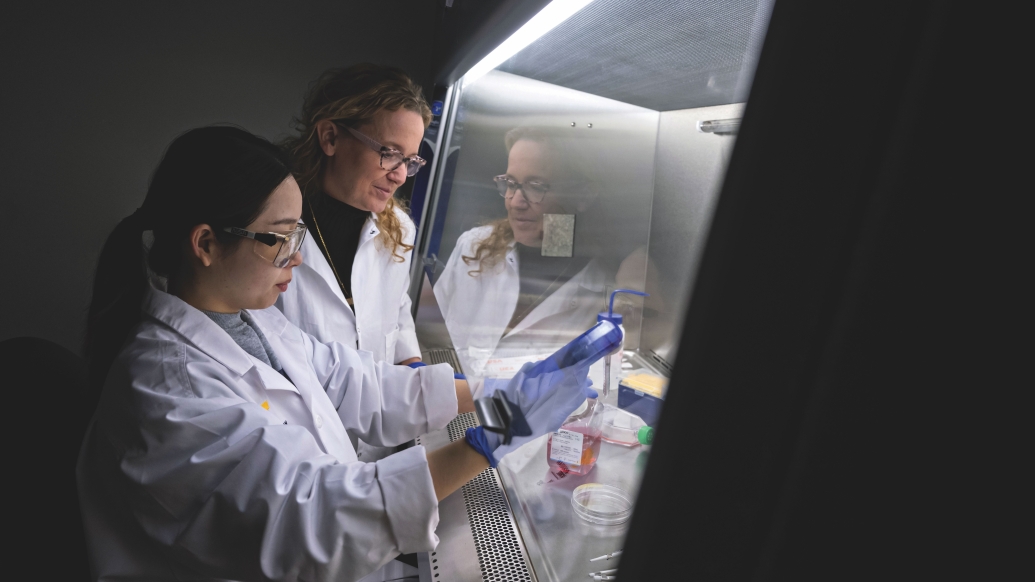 Michelle Hastings (right) and a student are working in the lab. They are wearing white lab coats and the student is holding lab equipment.