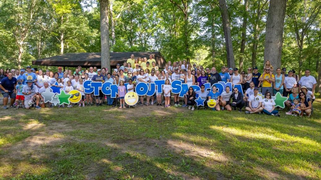 Group photo of roughly 100 people holding up letters that spell "Stomp Out Sarcoma" in a grassy, wooded area.