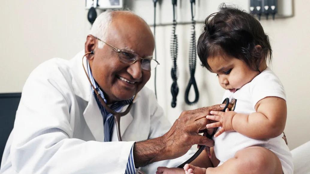 doctor close up listening to baby's heart while baby touches stethoscope while doctor smiles in doctor exam room on exam room table with medical equipment in background