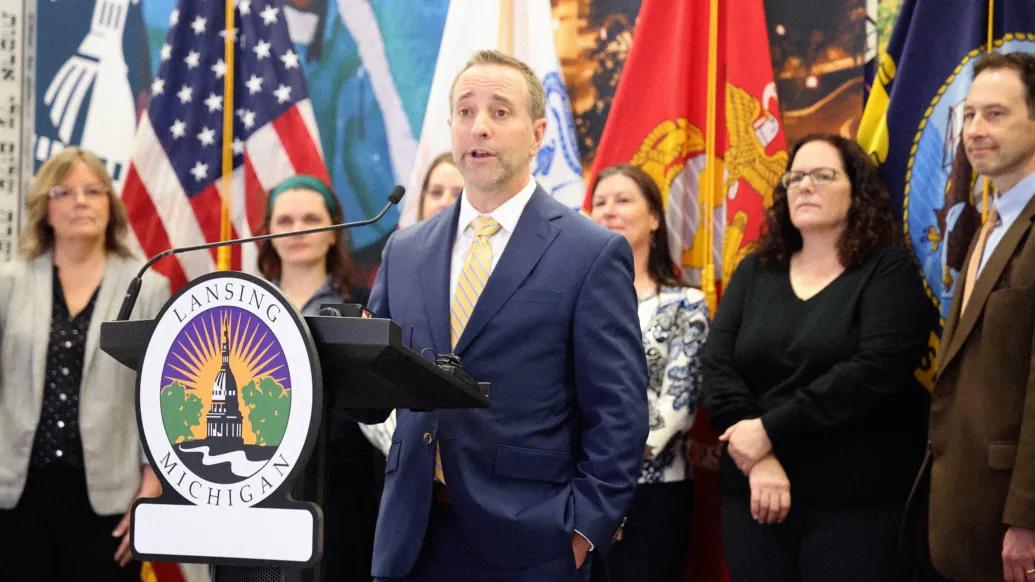 man speaking at podium with people standing behind him and flags