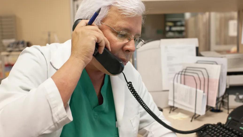 Doctor with white hair and white coat holding a phone to his ear.