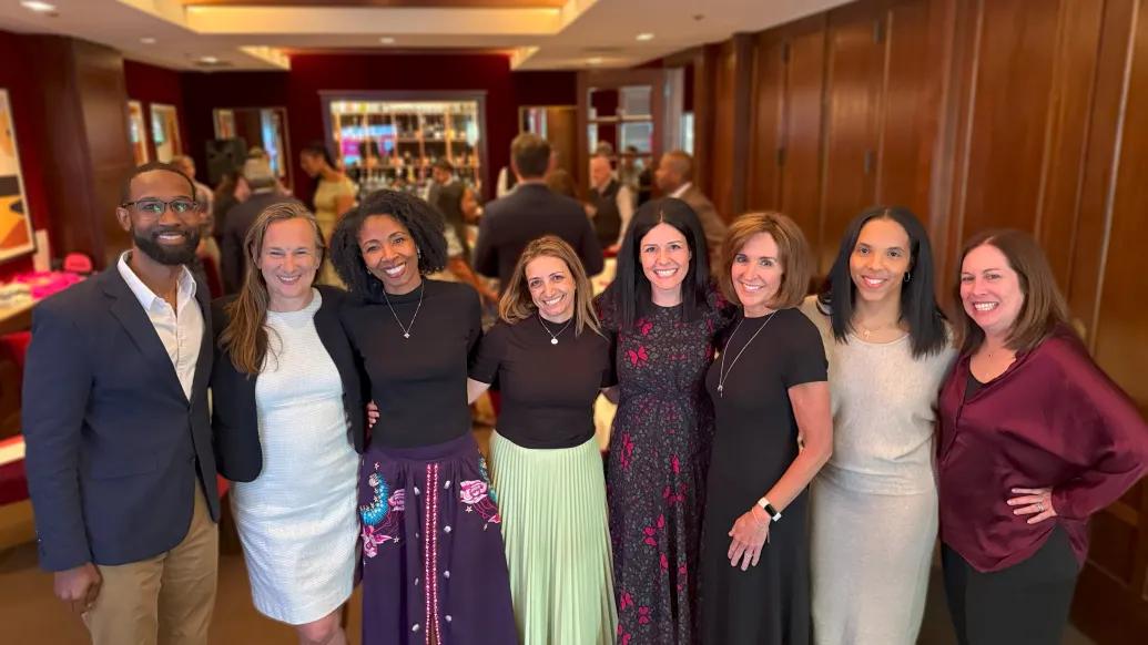 Smiling group shot of eight Bright Pink board members standing in a row together. They are wearing business casual and appear to be in the lobby of a restaurant.