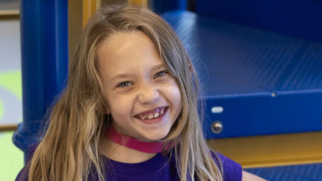Photo of Evalynn seated on the steps of a piece of play equipment. She has long brown hair and is wearing a purple Calming the Storm of Epilepsy t-shirt.