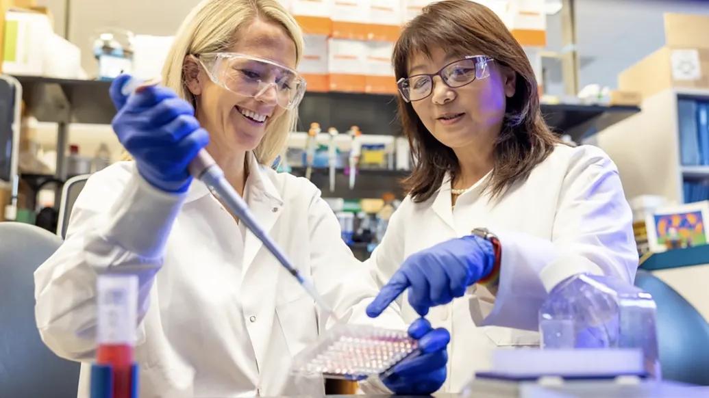 Two researchers working in a lab. They are smiling and wearing safety glasses, blue gloves, and white coats. One is using a pipette and the other points to what she is working on.