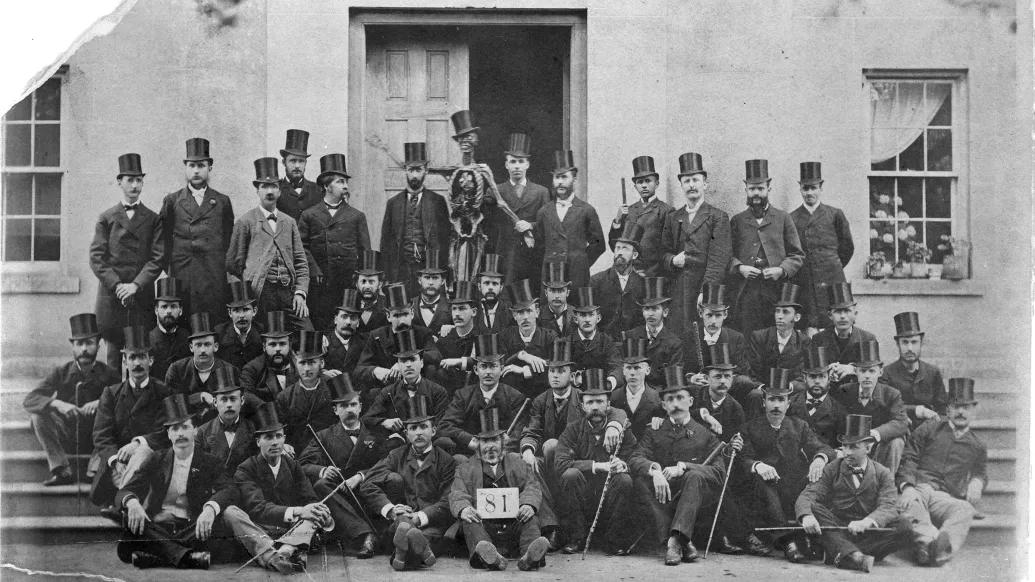 Group of Medical Students from 1881 wearing dark suits and top hats. In back, a student holds up a skeleton wearing a top hat.